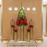Architectural Christmas tree installation in the First Class Lounge, featuring cascading Pinus greenery, red ornaments, and sculptural gold pedestals beneath a global time zone display.