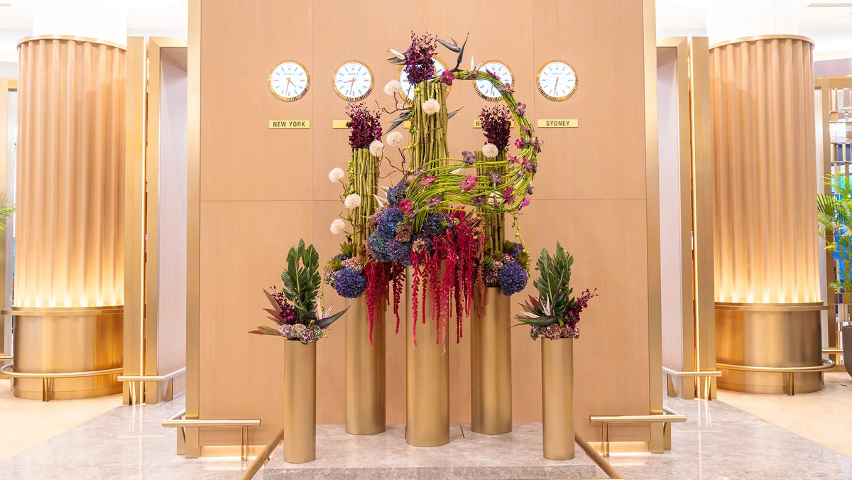 Luxury floral installation featuring cascading red amaranthus, purple hydrangeas, orchids, and birds of paradise on golden pedestals in an elegant lobby setting.