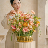 Woman arranging a pastel Easter floral arrangement in a gold vase, featuring soft peach and pink blooms with decorative eggs, set in a bright, neutral interior.