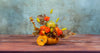 Autumn-themed floral arrangement in a small pumpkin vase, featuring orange roses, yellow accents, red berries, and dried oak leaves, displayed on a rustic wooden table against a textured blue-green background.