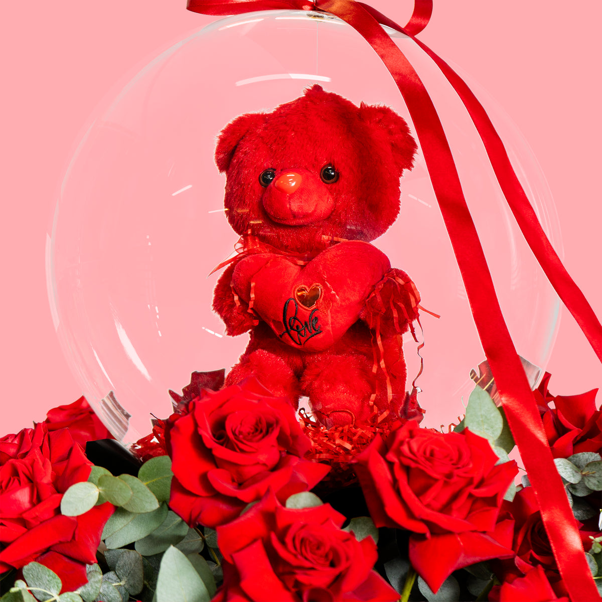 Red teddy bear with a heart, surrounded by red roses and a clear snow globe on a pink background