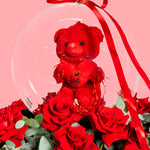 Red teddy bear with a heart, surrounded by red roses and a clear snow globe on a pink background
