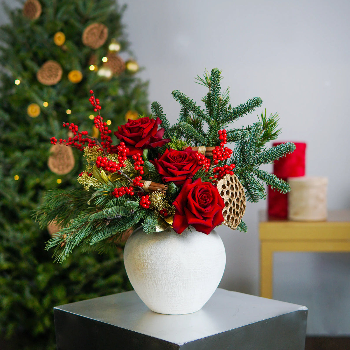 Decorative floral arrangement with red flowers and greenery in a white vase, Christmas tree in the background.