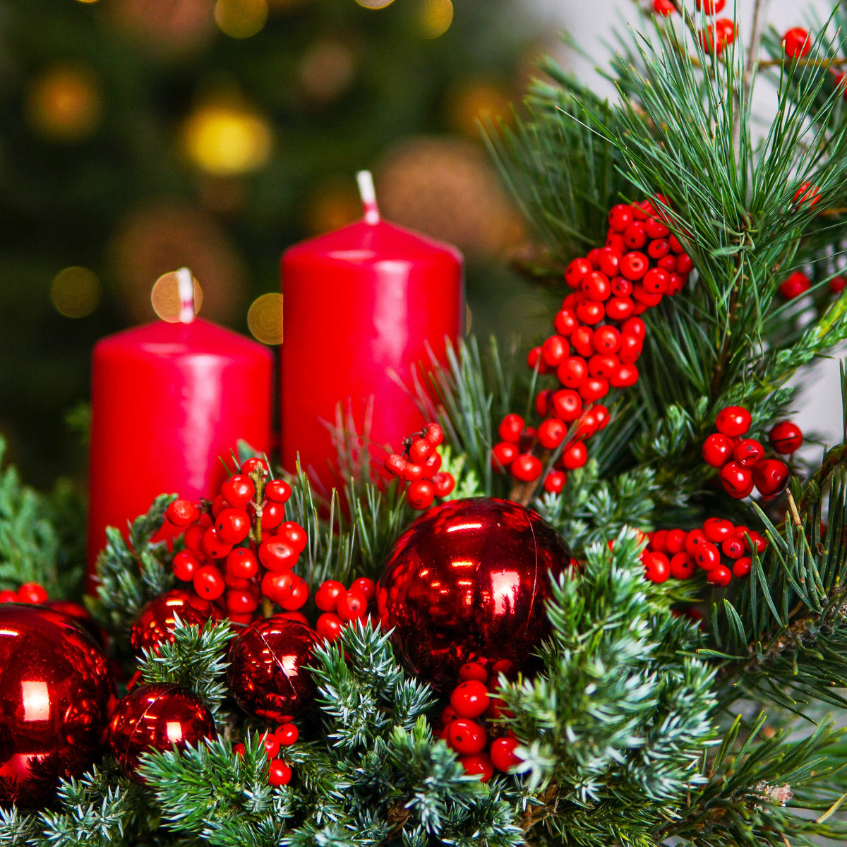 Decorative arrangement with red candles, berries, and ornaments on a blurred Christmas tree background.