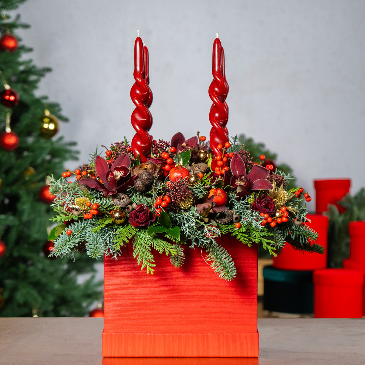 Decorative arrangement with red candles and flowers in an orange container against a Christmas tree background.