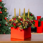 Decorative Christmas arrangement with gold candles, red flowers, and greenery in a red box, with a blurred Christmas tree in the background.