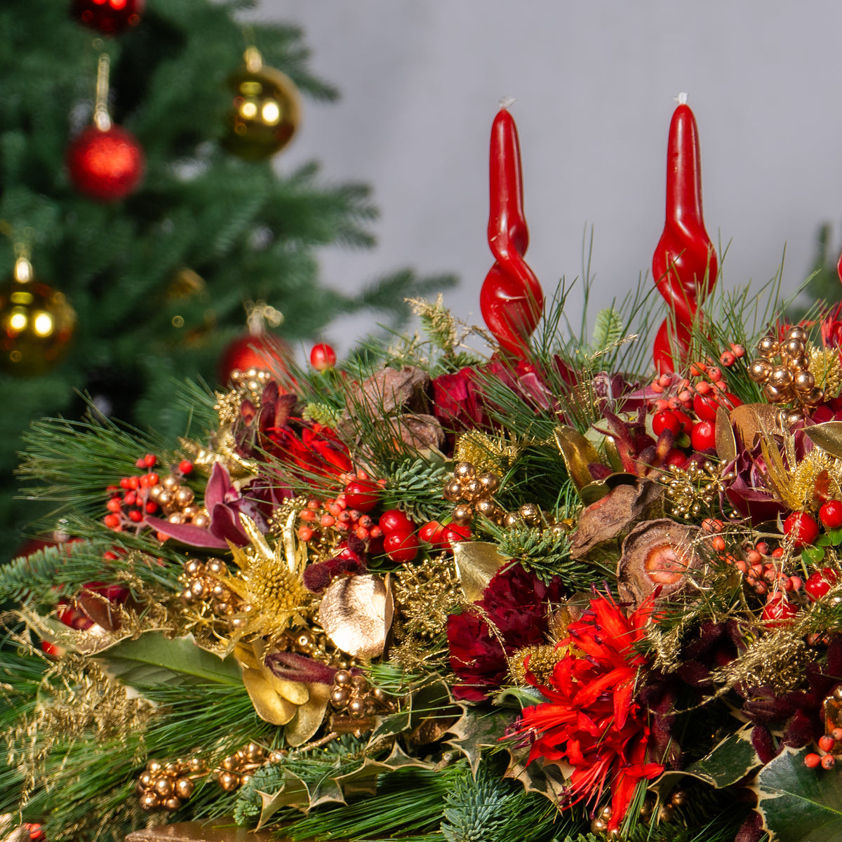 Decorative Christmas arrangement with red candles, gold ribbons, and greenery against a blurred Christmas tree background.