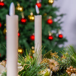 Two white candles in a festive arrangement with a blurred Christmas tree in the background.