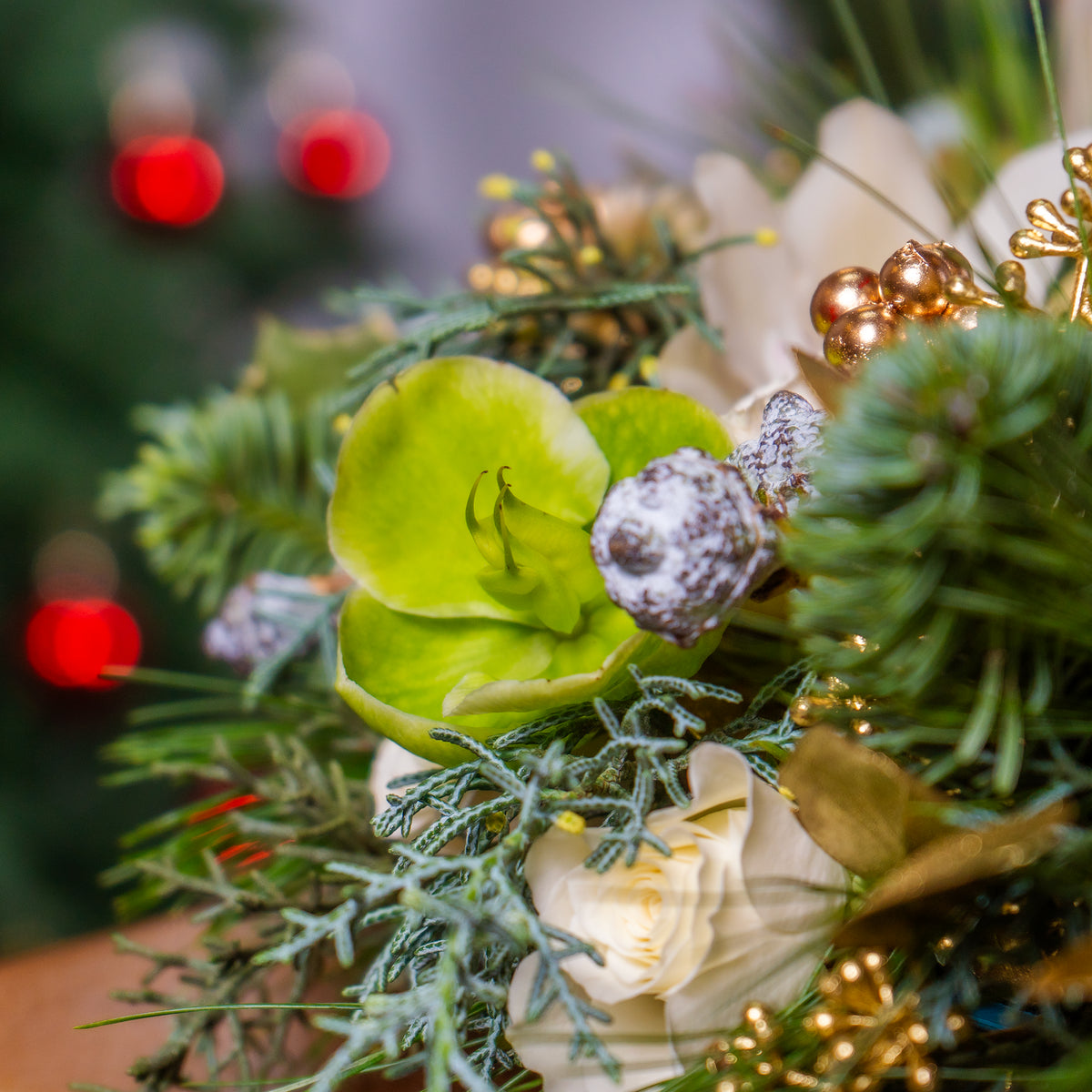 Close-up of a festive floral arrangement with greenery, white flowers, and gold accents against a blurred Christmas tree background.