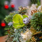 Close-up of a festive floral arrangement with greenery, white flowers, and gold accents against a blurred Christmas tree background.