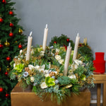 Decorative Christmas arrangement with candles and greenery on a table, with a Christmas tree in the background.