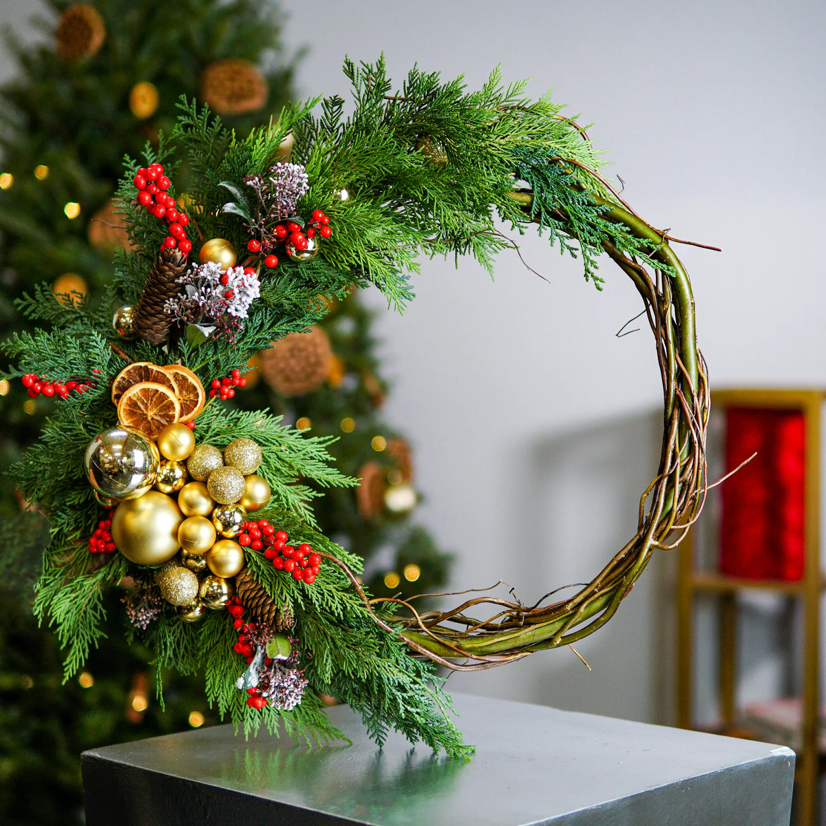 Decorative wreath with greenery, ornaments, and berries on a stand with a blurred Christmas tree in the background.
