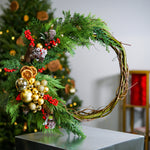Decorative wreath with greenery, ornaments, and berries on a stand with a blurred Christmas tree in the background.