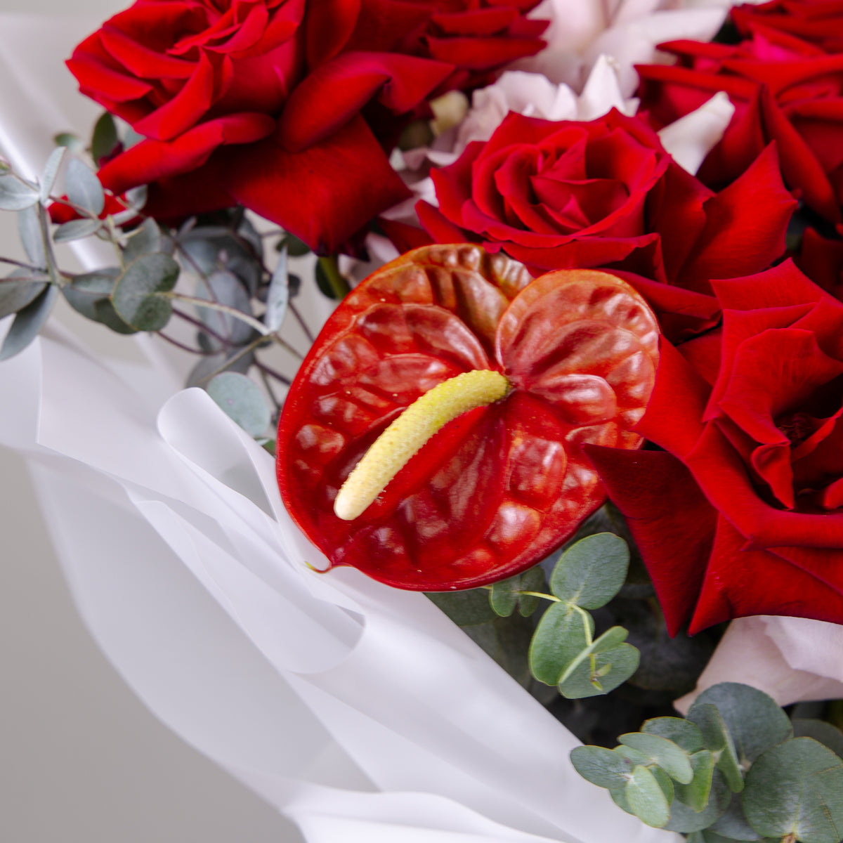Red anthurium flower with a yellow spadix nestled among red roses and eucalyptus leaves in the bouquet.