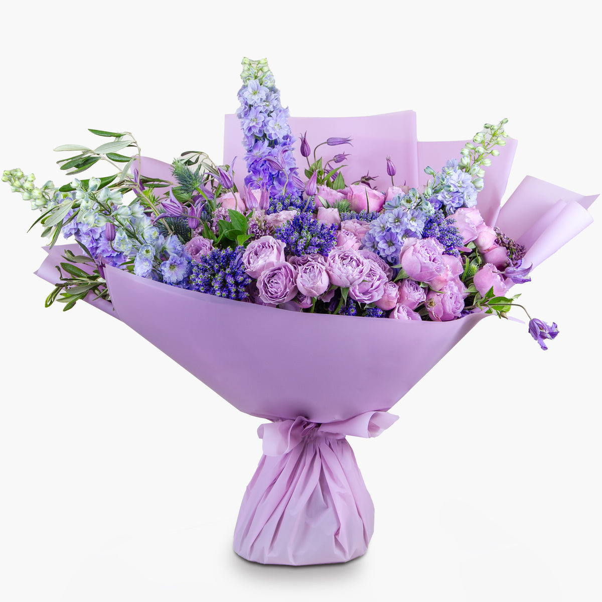 A bouquet of flowers featuring purple and lavender hues, including lilac roses, delphiniums, and veronica, wrapped in a matching lavender paper.