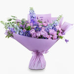 A bouquet of flowers featuring purple and lavender hues, including lilac roses, delphiniums, and veronica, wrapped in a matching lavender paper.