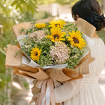 Bouquet of sunflowers and greenery wrapped in brown paper held by a person in a white dress.