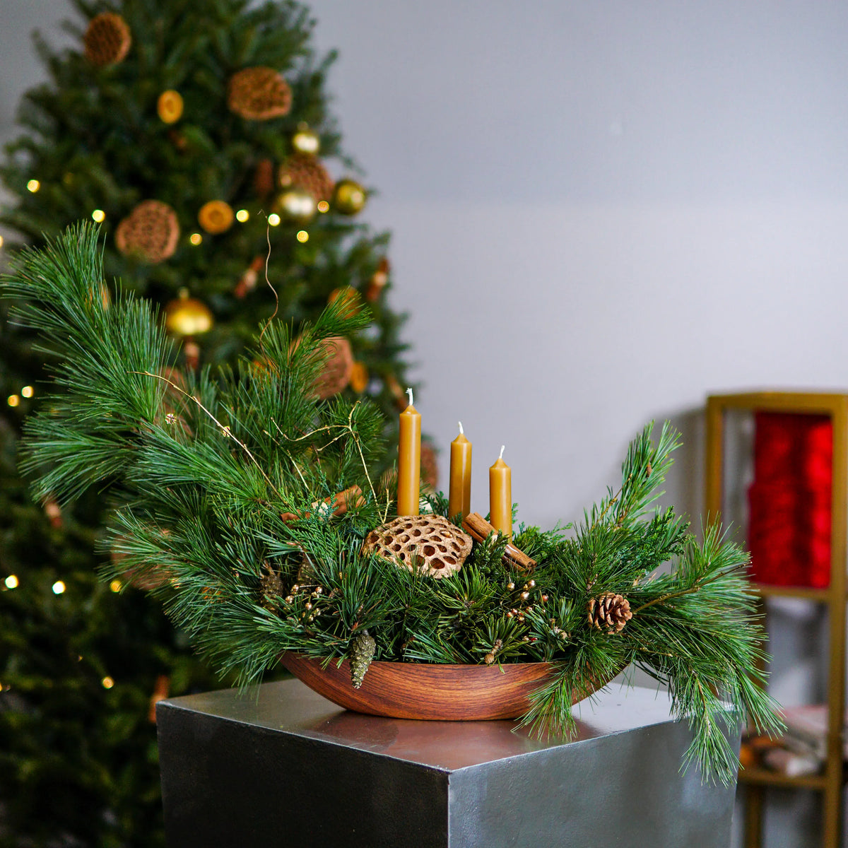 Decorative Christmas arrangement with candles and greenery in a wooden bowl, blurred Christmas tree in the background.
