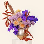 Colorful floral arrangement with purple and orange flowers in a metallic vase on a beige background