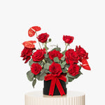Bouquet of red roses with greenery in a black vase on a white pedestal against a white background
