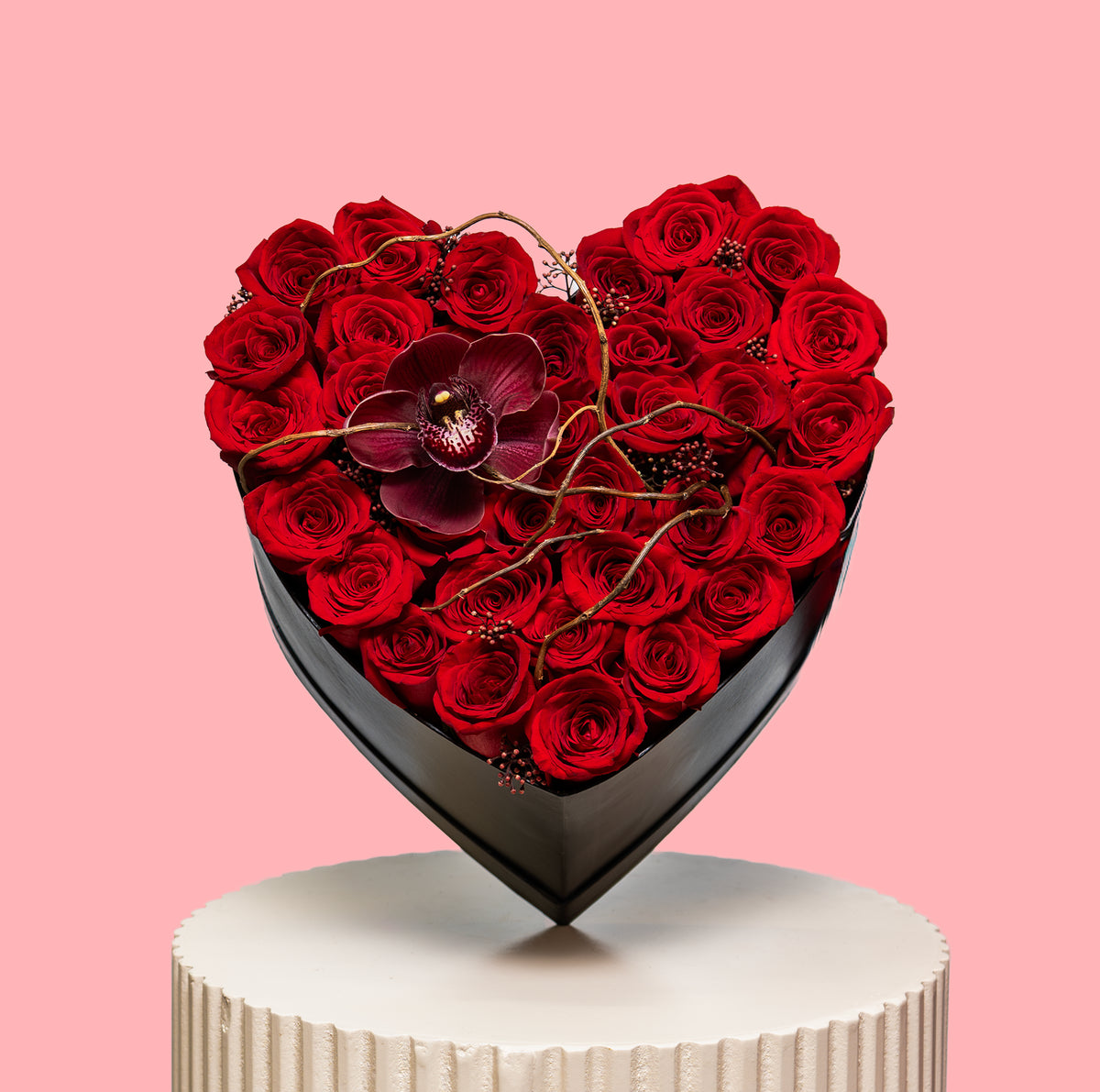 Heart-shaped arrangement of red roses with a central purple flower on a pink background