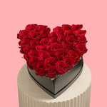 Heart-shaped arrangement of red roses in a black box on a white stand with a pink background