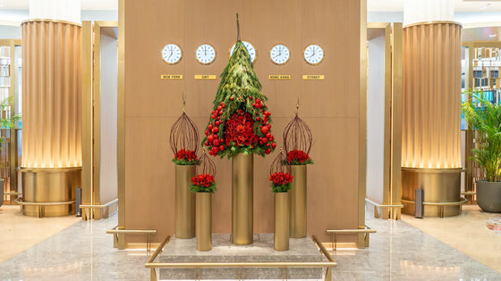 Architectural Christmas tree installation in the First Class Lounge, featuring cascading Pinus greenery, red ornaments, and sculptural gold pedestals beneath a global time zone display.