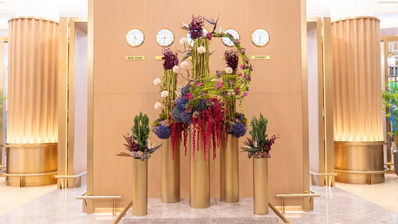 Luxury floral installation featuring cascading red amaranthus, purple hydrangeas, orchids, and birds of paradise on golden pedestals in an elegant lobby setting.