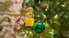 Close-up of Christmas tree ornaments featuring a miniature Scotch bottle decoration and a shiny green bauble, surrounded by warm string lights and evergreen branches.