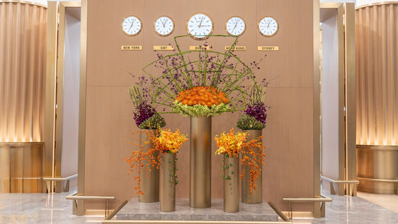 Large luxury floral installation with orange, green, and purple flowers arranged in tall gold vases under world clocks in a modern lobby.