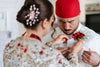Bride fixing a red floral boutonniere on the groom’s white attire, with intricate henna on her hands and jeweled hairpiece visible.