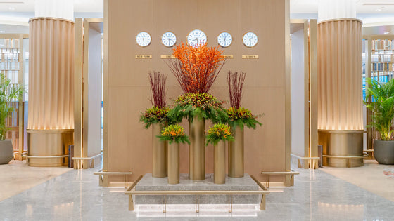 Tall gold vases filled with orange Ilex berries, red Salix branches, and mixed autumnal foliage arranged beneath world clocks in a luxury lounge.