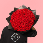 Bouquet of red roses held by a person wearing a black shirt with a logo on a pink background
