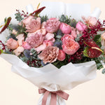 A large, vibrant flower bouquet featuring pink roses, peonies, thistles, and red anthuriums, wrapped in white paper with a dusty pink ribbon, set against a solid light beige background.