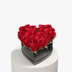 Heart-shaped box filled with red roses on a white pedestal against a white background
