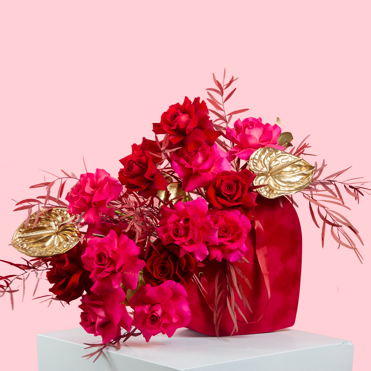 A vibrant flower arrangement featuring red roses, pink fuchsia, gold anthurium, and burgundy eucalyptus, presented in a heart-shaped box with a handle.