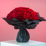 A bouquet of red roses wrapped in black paper with a ribbon tie against a pink background.
