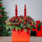 Decorative arrangement with red candles and flowers in an orange container against a Christmas tree background.