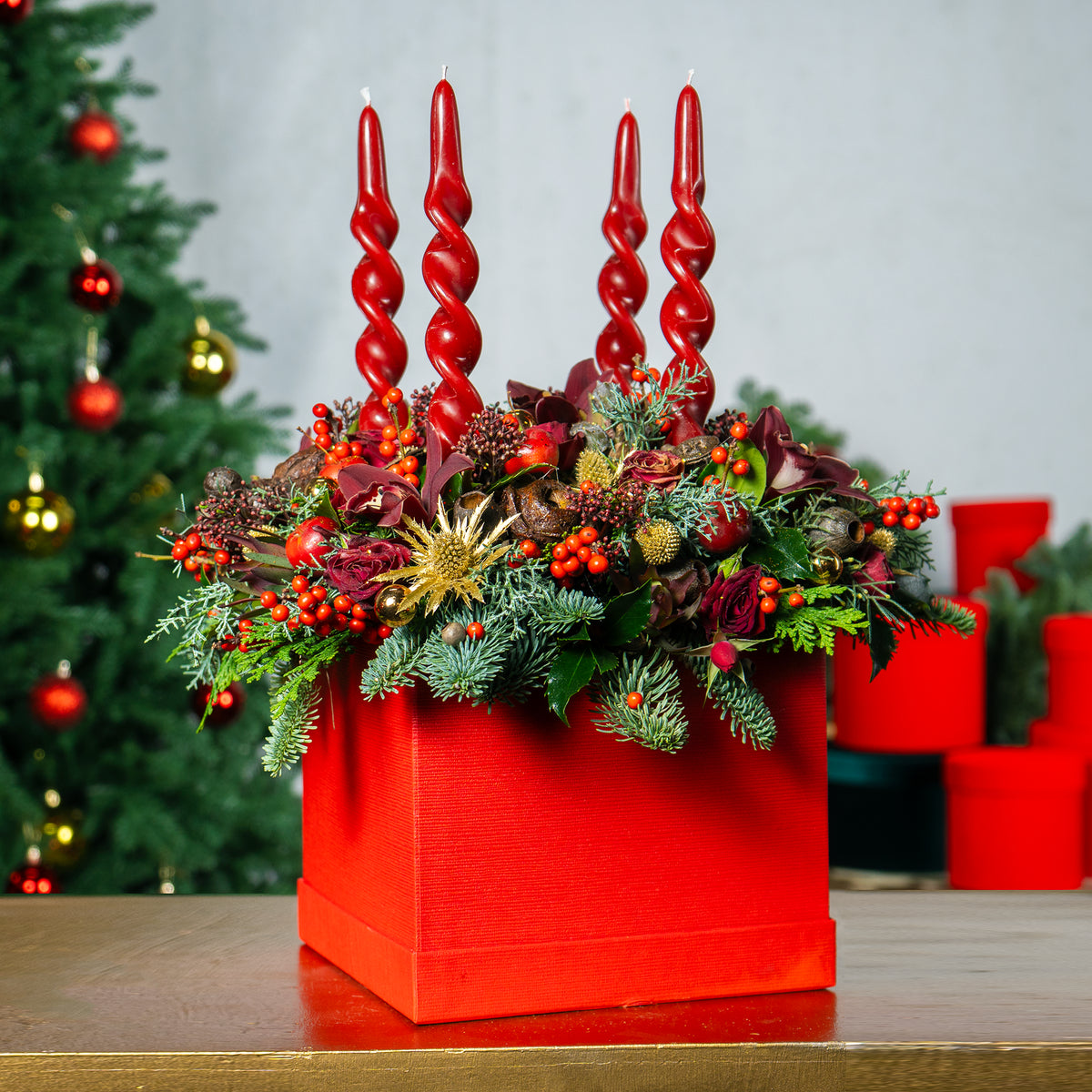 Decorative arrangement with red candles and festive elements on a table, Christmas tree in the background