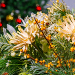 Close-up of a floral arrangement with yellow flowers and green leaves, blurred Christmas lights in the background.