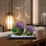 Decorative arrangement with purple flowers and green spherical elements on a dark base, set against a blurred indoor background.