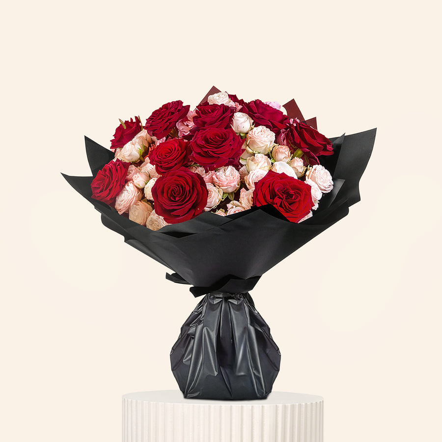 Bouquet of red and pink roses wrapped in black paper on a white pedestal against a light gray background