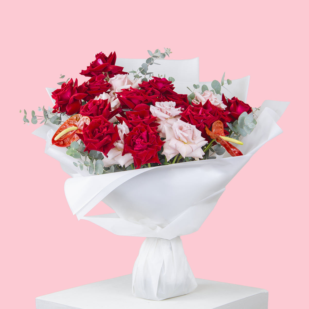 Elegant bouquet of red and pale pink roses with eucalyptus leaves, accented by red anthurium flowers, wrapped in crisp white paper against a pink background.