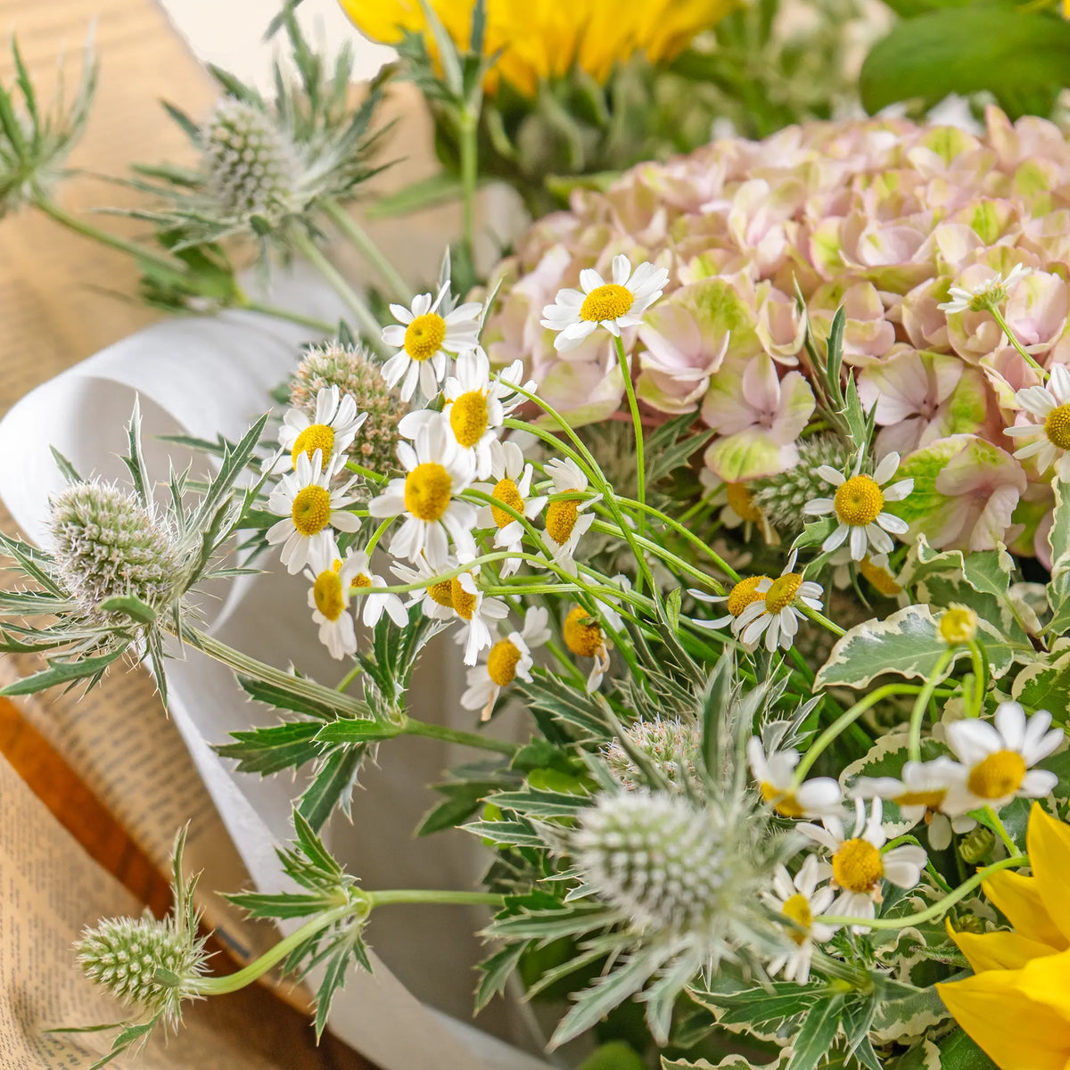 Bouquet of flowers with white and yellow daisies, greenery, and pink hydrangeas on a wooden surface.