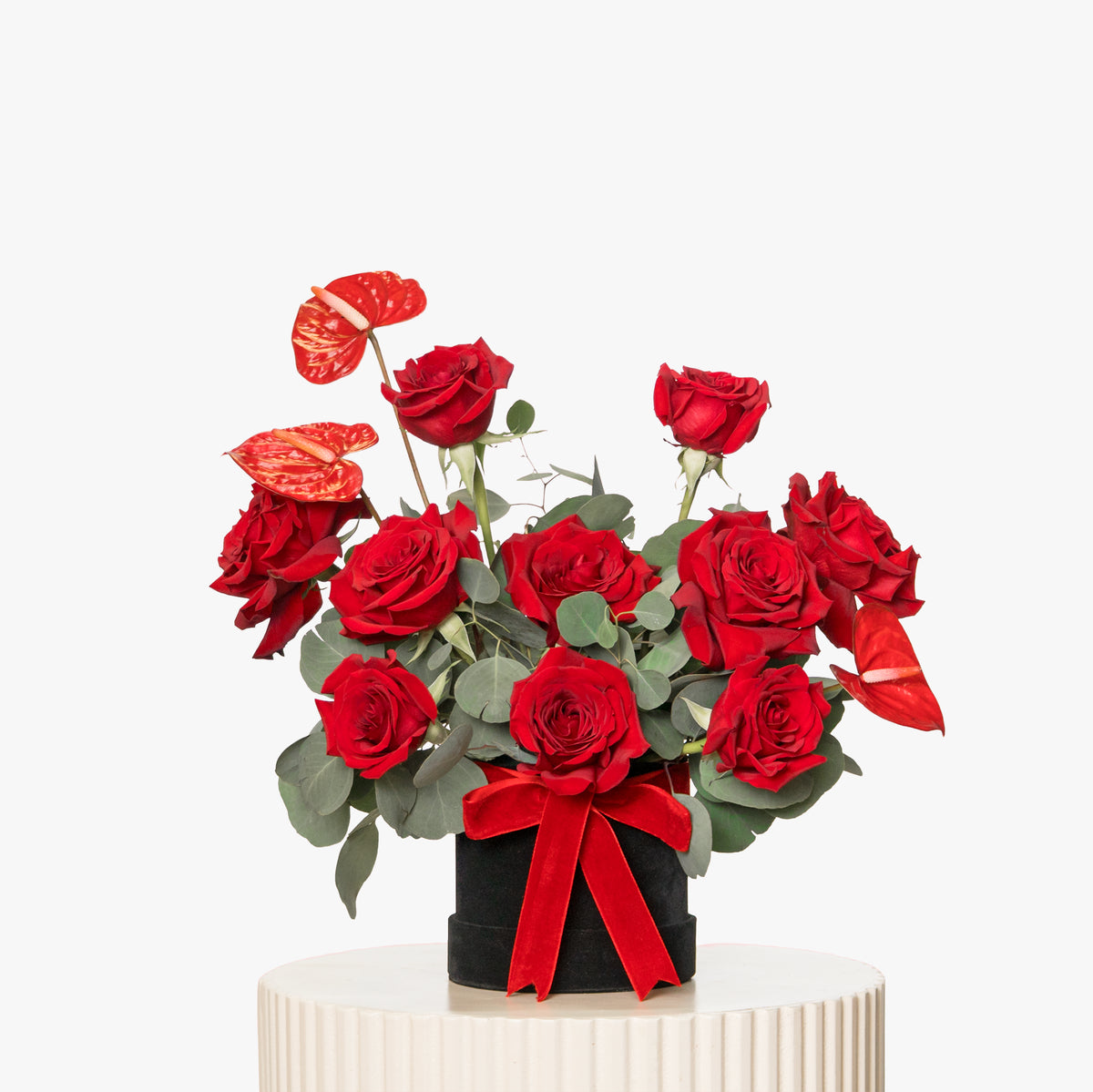 Bouquet of red roses with greenery in a black vase on a white pedestal against a white background