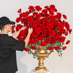 Person arranging red roses in a large gold vase against a pink background