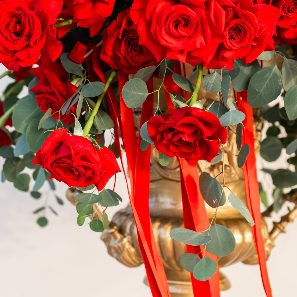 Bouquet of red roses with green leaves and red ribbons in a gold vase.