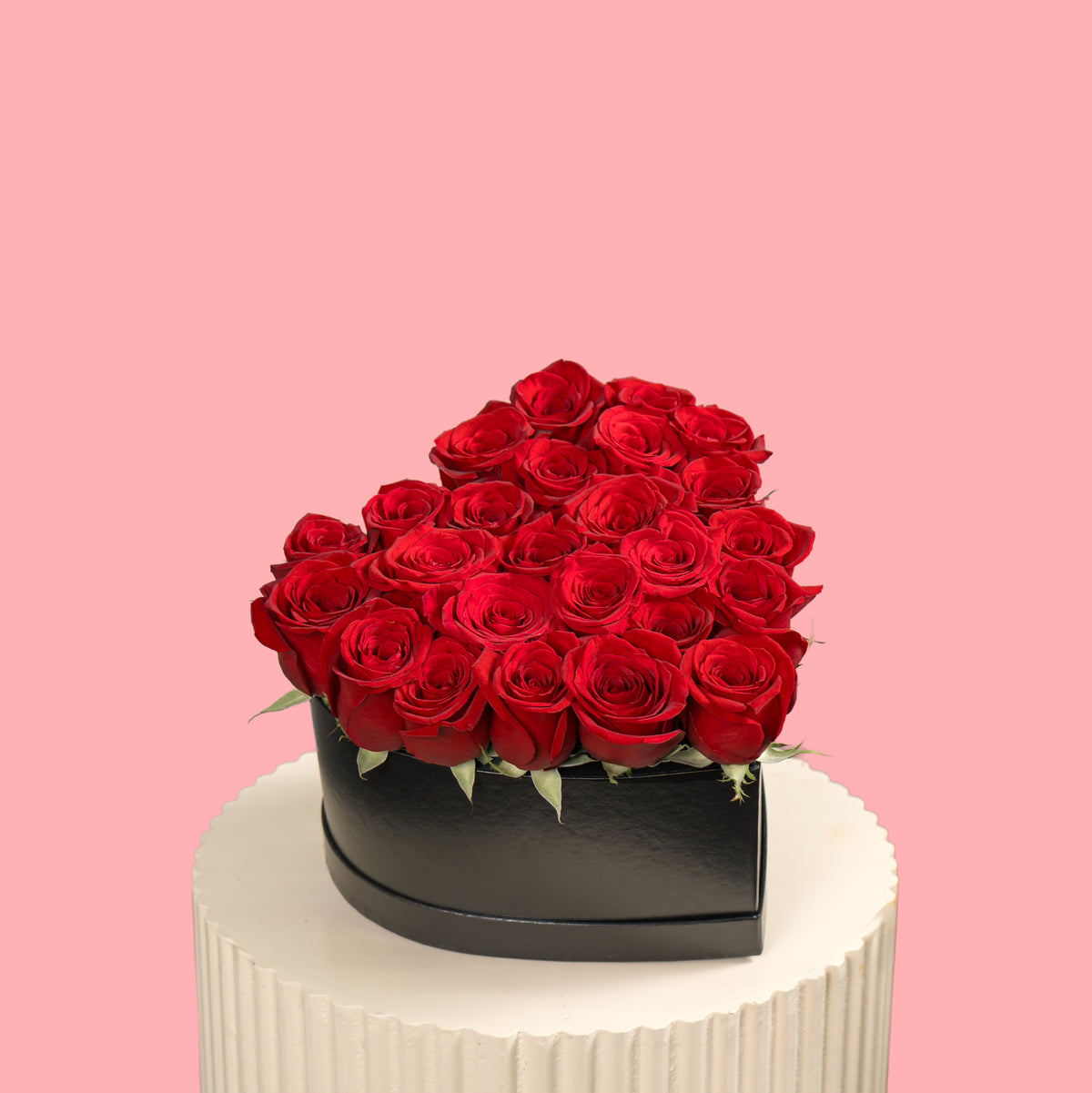 Heart-shaped arrangement of red roses in a black box on a white pedestal with a pink background