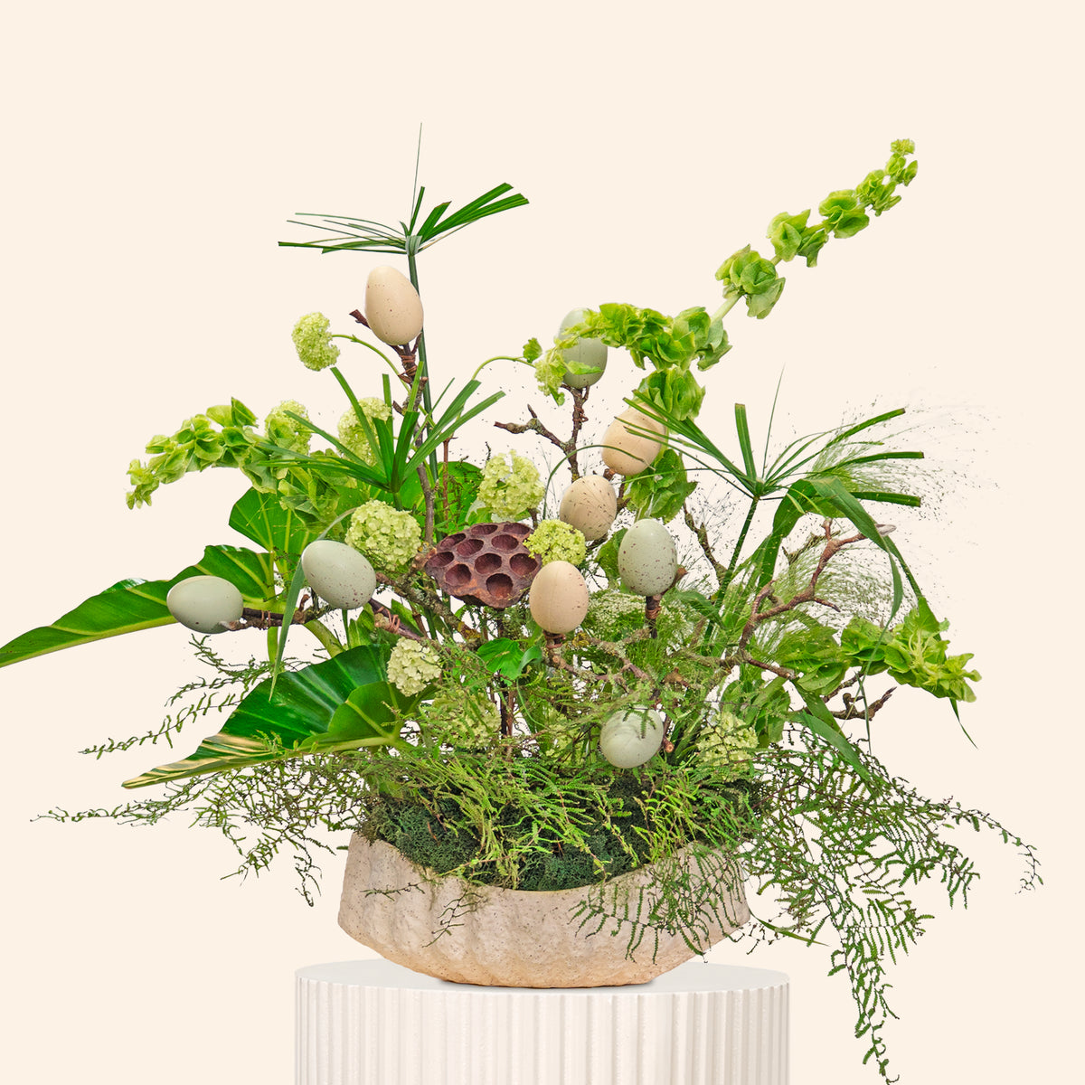Decorative plant arrangement with greenery and white eggs on a beige pedestal against a light beige background
