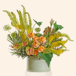 A round sage green floral arrangement in a matching round pot, featuring yellow-green feathery stems, orange roses, green leaves, and small dried botanicals, set against a plain light beige background.
